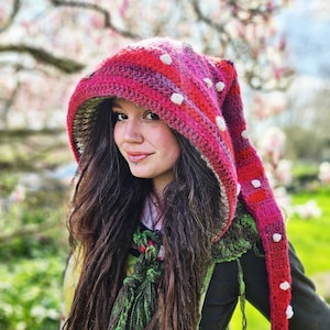 Op de afbeelding: Een vrouw met lange bruine dreadlocks draagt een rood-witte gebreide hoed in de vorm van een paddenstoel met witte stippen. Ze draagt een groen-bruin gepatroneerd topje en een zwart vest.