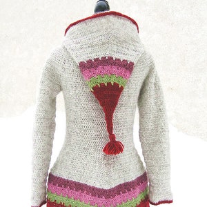 Può includere: Un cardigan a maglia con cappuccio, con un cappuccio lungo e appuntito e un bordo colorato. Il cardigan è realizzato in un colore beige chiaro con un bordo rosso, verde e rosa. Il cappuccio ha un fiocco all'estremità.