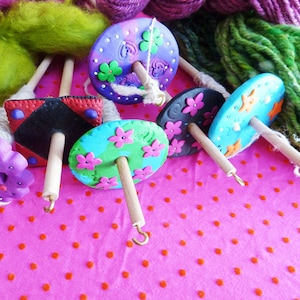 May include: Six colorful hand-painted pottery spinning wheels with wooden spindles and metal hooks. The wheels are decorated with flowers and other designs. The wheels are on a pink background with white polka dots.