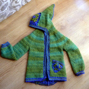Puede incluir: Chaqueta de punto de ganchillo con capucha a rayas verdes y azules con un diseño floral en la capucha y un bolsillo en la parte delantera.
