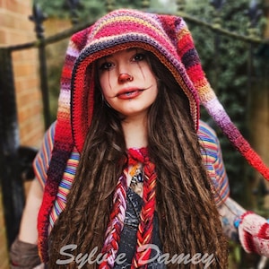 Peut inclure: Une femme aux longs dreadlocks porte une écharpe à capuche en crochet colorée avec une longue pointe pointue. L'écharpe est rayée de rouge, orange, jaune, vert, bleu, violet et noir. La femme porte une chemise rayée et a une écharpe tressée colorée autour du cou.