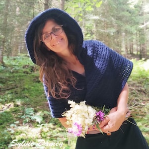 Peut inclure: Une femme portant un châle en crochet à capuche bleu foncé avec un bouquet de fleurs blanches dans les mains. Elle se tient dans un environnement forestier.