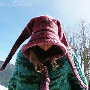 May include: A woman wearing a purple and green crocheted hooded sweater with long, pointed ear-like extensions. The sweater has a striped pattern and the hood is pulled down over her head.