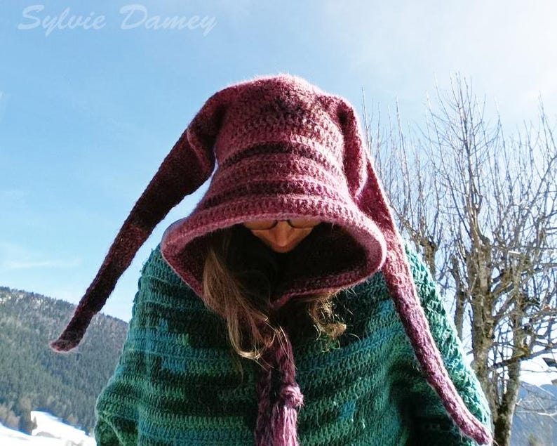 May include: A woman wearing a purple and green crocheted hooded sweater with long, pointed ear-like extensions. The sweater has a striped pattern and the hood is pulled down over her head.