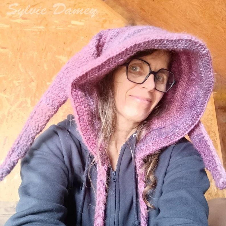 May include: A woman wearing a purple and pink hooded hat with long ear-like flaps. The hat is crocheted and has a textured look. The woman is wearing glasses and has long brown hair.