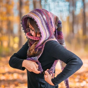May include: A woman wearing a multi-colored crochet hooded scarf with a long, flowing tail. The scarf is made with a variety of shades of purple, pink, brown, and beige yarn. The woman is standing in a forest setting with fall foliage in the background.