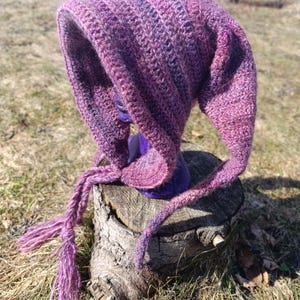 May include: A purple and pink crocheted hooded hat with a long tassel. The hat is sitting on a tree stump.