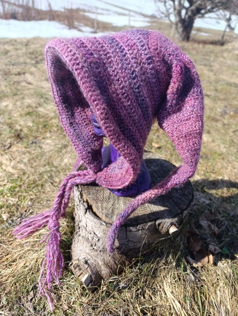May include: A purple and pink crocheted hooded hat with a long tassel. The hat is sitting on a tree stump.