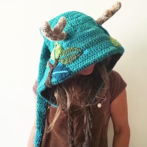 Puede incluir: Un gorro de punto azul turquesa con cuernos marrones y aplicaciones de hojas verdes. El gorro tiene una cola larga de punto.
