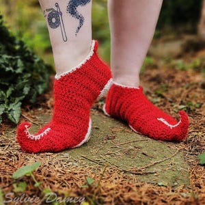 May include: Red crocheted elf shoes with white trim. The shoes have pointed toes and a curled up point at the heel. The shoes are on a bed of brown pine needles.