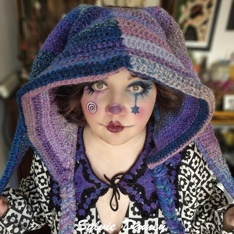May include: A person wearing a purple and blue crocheted hooded scarf with a black and white patterned shirt. The person has clown makeup on their face with a spiral, star, and teardrop design.