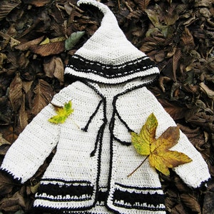 May include: A white crocheted hooded cardigan with black trim and a pointed hood. The cardigan is lying on a bed of brown leaves.