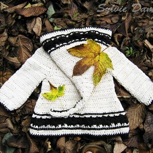 Op de afbeelding: Een wit gebreid babyvestje met zwarte bies en een contrasterende zwarte gebreide kraag. Het vestje is versierd met twee gele en bruine bladeren.