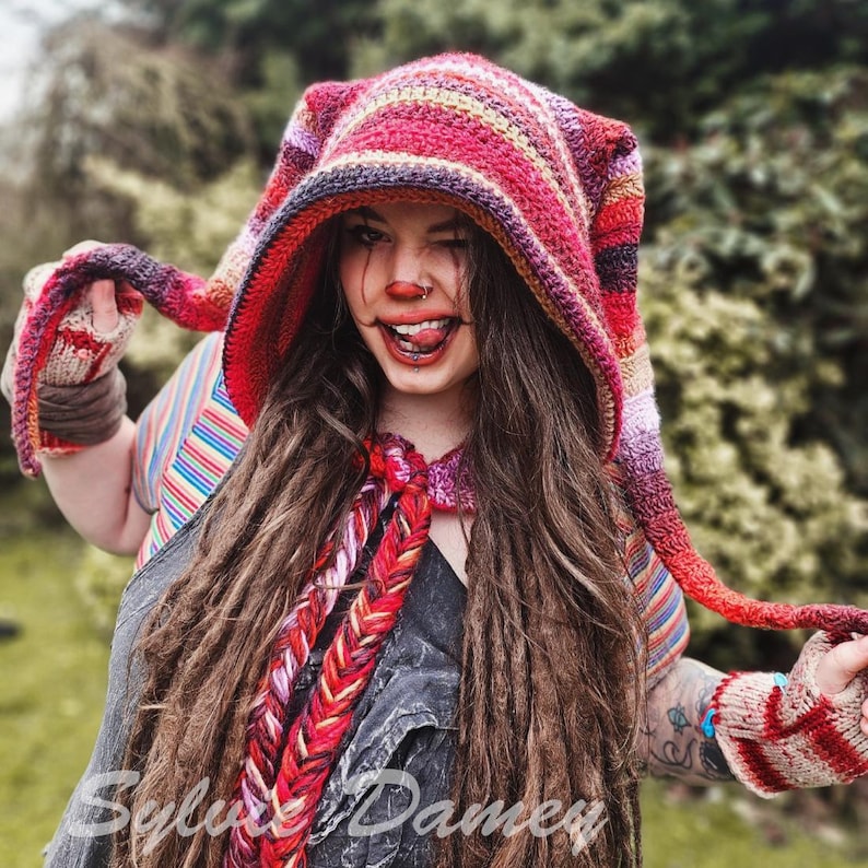 May include: A woman wearing a red, white, and brown striped hooded crocheted hat with long brown dreadlocks. The hat has long, dangling ties. She is wearing a striped shirt and a black shirt underneath.