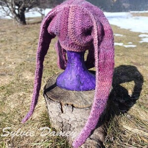 May include: A purple and pink knitted hat with long ear flaps. The hat is displayed on a purple mannequin head.