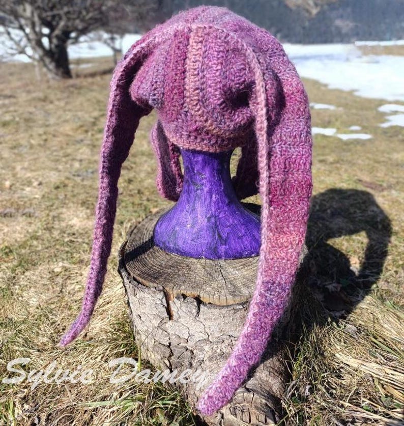 May include: A purple and pink knitted hat with long ear flaps. The hat is displayed on a purple mannequin head.