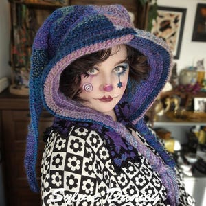 May include: A person wearing a purple and blue crocheted hooded scarf with a black and white patterned shirt. The person has clown makeup on their face.