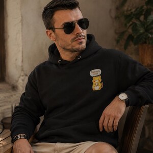 Puede incluir: Una sudadera negra con capucha que presenta un pequeño gráfico bordado en el pecho izquierdo de un tigre amarillo con un bocadillo que contiene el texto Namaste. La sudadera tiene mangas largas, un bolsillo canguro y capucha con cordón.