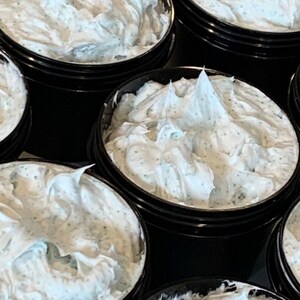 May include: Several open black jars filled with a white, creamy product. The product has a textured appearance with small, dark green specks throughout. The jars are arranged closely together, with the product overflowing slightly from each container.