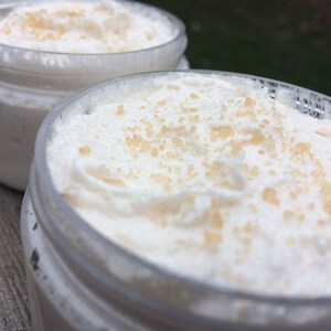 May include: Two clear jars filled with a white, whipped cream-like substance, speckled with light brown granules. The jars are partially visible, with the focus on the creamy contents. The background is blurred.