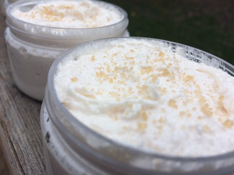 May include: Two clear jars filled with a white, whipped cream-like substance, speckled with light brown granules. The jars are partially visible, with the focus on the creamy contents. The background is blurred.