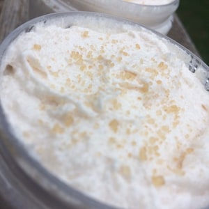 May include: Close-up of two clear jars filled with a white, whipped cream-like substance. The cream is topped with small, light brown granules. The jars are partially visible, with a blurred background.