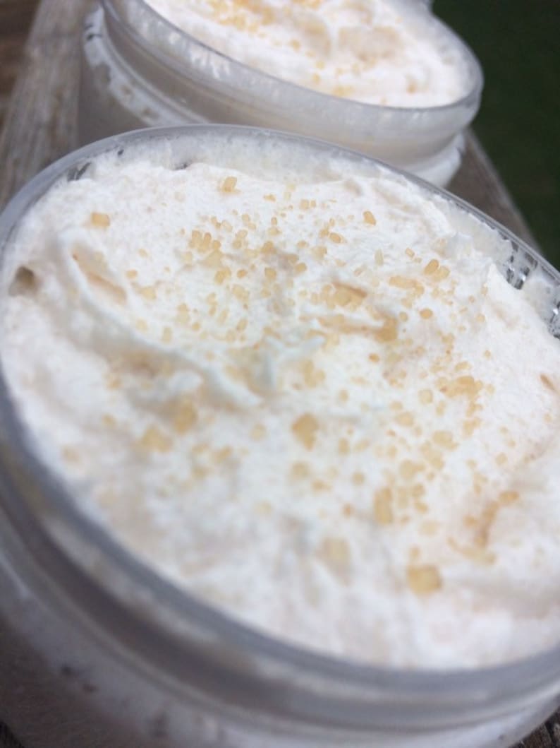 May include: Close-up of two clear jars filled with a white, whipped cream-like substance. The cream is topped with small, light brown granules. The jars are partially visible, with a blurred background.