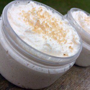May include: Close-up of three clear plastic jars filled with a white, whipped cream-like substance. The substance is topped with small, light brown granules. The jars are arranged in a row, with the focus on the first jar.