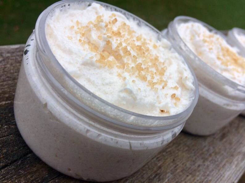 May include: Close-up of three clear plastic jars filled with a white, whipped cream-like substance. The substance is topped with small, light brown granules. The jars are arranged in a row, with the focus on the first jar.
