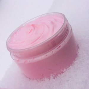 May include: A clear, round jar filled with a light pink, whipped cream-like substance. The product has a smooth, swirled texture and is contained in a transparent plastic jar. The jar is sitting on a white surface.
