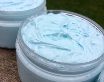 May include: Close-up of three clear jars filled with light blue, whipped cream-like substance. The jars are sitting on a wooden surface, with a blurred green background. The product appears to be a cosmetic or skincare item.