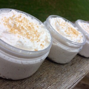 May include: Several small, clear jars filled with a creamy, white substance, topped with a scattering of light brown granules. The jars are arranged in a row on a wooden surface, suggesting a skincare or cosmetic product.