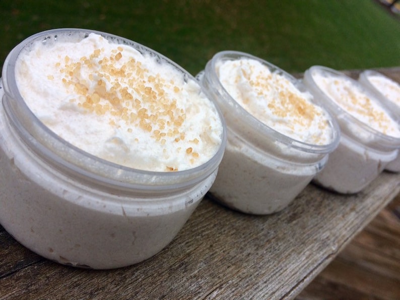 May include: Several small, clear jars filled with a creamy, white substance, topped with a scattering of light brown granules. The jars are arranged in a row on a wooden surface, suggesting a skincare or cosmetic product.