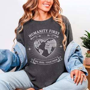 Może przedstawiać: Ciemnoszara koszulka z napisem "HUMANITY FIRST" nad grafiką mapy świata w kształcie serca. Poniżej grafiki znajduje się napis "WE RISE TOGETHER". Koszulka jest noszona z dżinsową kurtką.