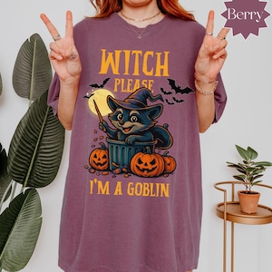 Op de afbeelding: Bessenkleurig T-shirt met een Halloween-afbeelding. Het ontwerp toont een wasbeer met een heksenhoed, die een toverstaf vasthoudt, met pompoenen en de tekst "Witch Please I'm a Goblin". Vleermuizen en een volle maan zijn ook inbegrepen.