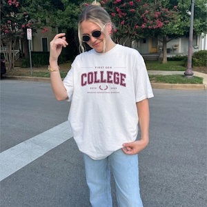 May include: A white t-shirt featuring the text "FIRST GEN COLLEGE" in burgundy. The shirt also displays "ESTD 2023" and "BREAKING GENERATIONAL BARRIERS". The model is wearing light blue jeans and sunglasses.