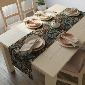 May include: A dining table set for a meal, featuring a floral patterned table runner with plates, napkins, and a small vase. The plates are beige and peach, and the napkins are folded. The table is made of light-colored wood.