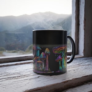 May include: Black ceramic mug featuring a colorful illustration of psychedelic mushrooms. The mushrooms are depicted in vibrant hues of blue, green, purple, and yellow. The mug is set against a window with a mountain view, and steam rises from the mug.