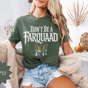 Model sitting in a cozy room wearing a moss green unisex graphic tee with “Don’t Be a Farquaad” text and crown stool illustration. Perfect funny shirt for meme fans and sarcastic gifts.