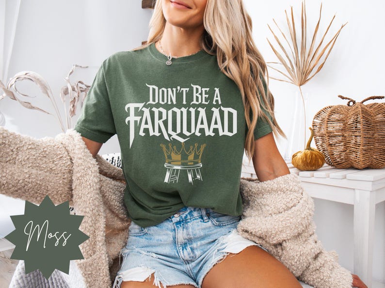 Model sitting in a cozy room wearing a moss green unisex graphic tee with “Don’t Be a Farquaad” text and crown stool illustration. Perfect funny shirt for meme fans and sarcastic gifts.