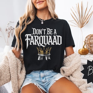 Woman wearing a black “Don’t Be a Farquaad” t-shirt with medieval-style text and a gold crown on a stool design. Meme-inspired graphic tee for Shrek fans and streetwear lovers. Trendy fall home setting.