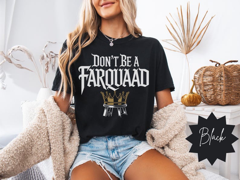 Woman wearing a black “Don’t Be a Farquaad” t-shirt with medieval-style text and a gold crown on a stool design. Meme-inspired graphic tee for Shrek fans and streetwear lovers. Trendy fall home setting.
