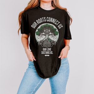 Può includere: T-shirt nero con grafica di un albero all'interno di un cerchio. Il testo "OUR ROOTS CONNECT US" è sopra l'albero e "OUR LOVE SUSTAINS US" è sotto. Tre piccoli cuori sono nella parte inferiore del disegno. La maglietta è indossata con jeans blu.