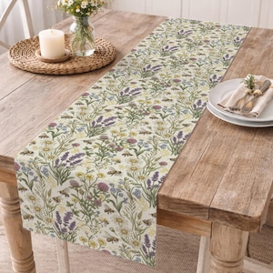 Puede incluir: Un camino de mesa rectangular con un estampado floral que presenta lavanda, margaritas y abejas. El camino de mesa está colocado sobre una mesa de madera, con una vela, flores y platos. La paleta de colores incluye crema, morado, amarillo y verde.