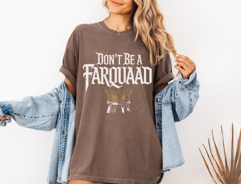 May include: Brown t-shirt with the text "DON'T BE A FARQUAAD" in a stylized font, with a crown graphic. The shirt has rolled-up sleeves and a relaxed fit.