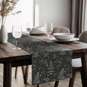 May include: A black table runner with a botanical design featuring ferns and wildflowers in beige and white. The runner is on a dark wooden table, with wine glasses, plates, and a vase visible.