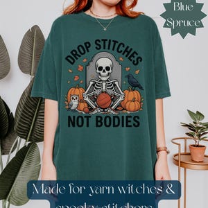 May include: Teal t-shirt with a Halloween graphic. The design shows a skeleton knitting with pumpkins, an owl, and a raven. Text reads "Drop Stitches Not Bodies" and "Made for yarn witches & spooky stitchers."