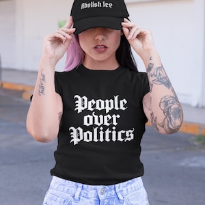 以下が含まれることがあります： 白い文字で「Abolish Ice」と書かれた黒い野球帽。ゴシック体のフォントで「People over Politics」という言葉が書かれた黒いTシャツを着ています。青いジーンズも履いています。