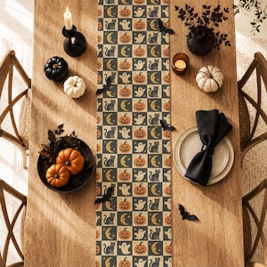 May include: A rectangular table runner featuring a repeating grid pattern of orange pumpkins, white ghosts, black cats, and yellow crescent moons on a dark background, displayed on a wooden table with decorative pumpkins and black napkins.