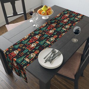 May include: A festive table runner featuring a repeating pattern of nutcrackers and Christmas trees on a dark background. The runner is placed on a dark wooden table, with plates, silverware, and wine glasses. A bowl of fruit is also present.